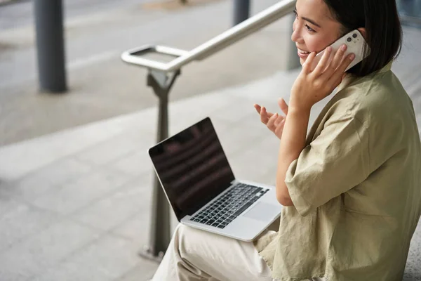 stock image Smiling asian woman makes a phone call. Girl student using laptop and mobile phone, talking to someone on telephone.