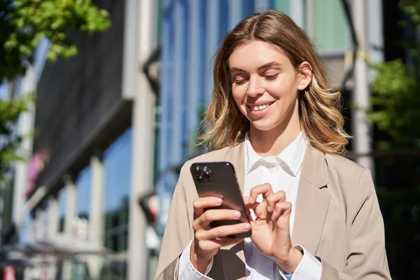 stock image Portrait of smiling beautiful businesswoman using her mobile phone on street. Corporate woman tap her smartphone, messaging, standing on street.