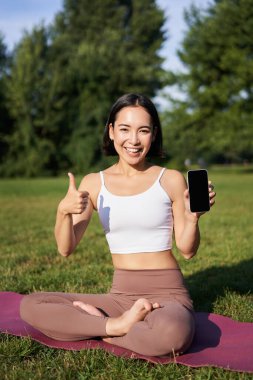 Asyalı bir kadının dikey çekimi, başparmaklarını gösteriyor, internetten yoga eğitimi öneriyor, meditasyon uygulaması yapıyor, parkta temiz hava egzersizleri yapıyor, akıllı telefon ekranını gösteriyor..