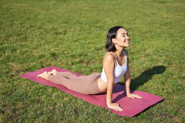 Gülümseyen genç sporcu parkta lastik mindere uzanıyor, yoga yapıyor, spor yapıyor, spor kıyafetleriyle temiz hava alıyor..