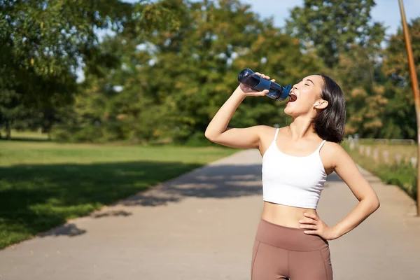 Stock image Happy asian sportswoman, runner drinks water from bottle while running, workout on fresh air in park.