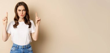 Frustrated young woman pointing fingers up, frowning displeased, standing in white t-shirt over beige background.