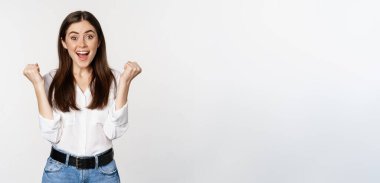 Excited young adult woman reacting to win, surprise news, screaming and cheering, triumphing, achieve goal and celebrating, standing over white background.