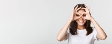 Gülümseyen, gülümseyen esmer kızın yakın plan fotoğrafı. Tamam jestler, el gözlükleri, beyaz arka plan..