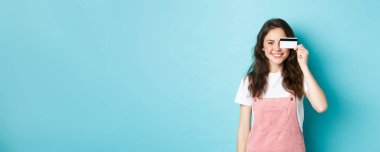 Gözlerinde plastik kredi kartı gösteren, gülümseyen, onunla alışveriş yapan, mavi arka planda duran hoş bir genç kadın..