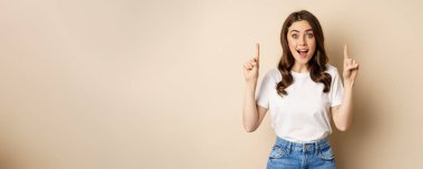 Enthusiastic young woman, female customer pointing fingers up and smiling, showing banner or logo, standing against beige background.