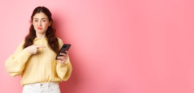 Şüpheci genç kadın tatminsiz suratını buruşturuyor, akıllı telefon ekranını işaret ediyor, online içerik hakkında şüpheleri var, pembe arka planda duruyor..