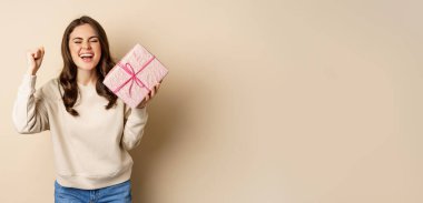 Happy smiling woman holding gift and triumphing, raising hand up joyfully, receive present on holiday, standing over beige background.