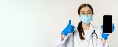 Online medical help concept. Enthusiastic young woman doctor in face mask, showing thumbs up and mobile phone app, smartphone screen interface, white background.