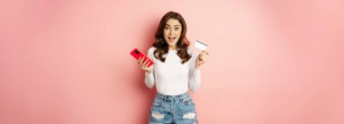 Enthusiastic brunette girl showing credit card and using mobile phone to order or pay, online shopping app, standing over pink background.