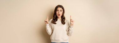 Amazed girl checking out banner announcement, reading promo text upwards, pointing fingers up, standing over beige background.