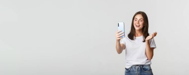 Arkadaş canlısı, çekici bir kız arkadaş, gülümseyen ve sohbet eden, akıllı telefon tutan, beyaz arka plan.