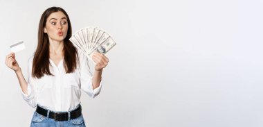 Excited woman holding credit card and money, looking amazed at cash, standing against white background. Copy space