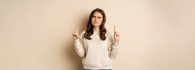Confused young woman pointing fingers up and staring puzzled, cant understand promo text upwards, beige background.