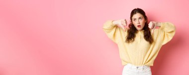 Ne kadar aptalca. Hoşnutsuz ve ilgisiz genç kadın gözlerini devirmiş, iç çekiyor ve yan bakıyor, şüpheli yüzüyle baş parmaklarını gösteriyor, pembe arka planda duruyor..