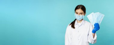 Portrait of medical worker, woman doctor in suggesting face masks from covid-19 pandemic, standing in hospital uniform over torquoise background.