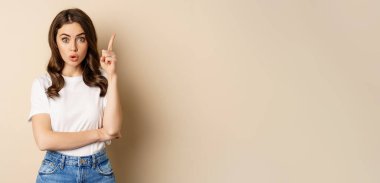 Excited woman raising finger, pitching an idea, has suggestion, wearing white t-shirt and jeans over beige background.