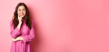 Heyecanlı güzel bir kız gülümsüyor, parmağını ısırıyor ve cezbedici bir yüzle kameraya bakıyor, ilginç bir fikre sahip, ilgi çekici bir şeye bakıyor, pembe arka planda duruyor..