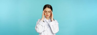 Image of woman doctor, medical personel grimacing from discomfort, having headache, migraine, feeling deezy, standing over torquoise background.
