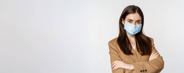 Coronavirus and pandemic concept. Professional businesswoman, office worker in medical face mask cross arms on chest, looking confident, white background.