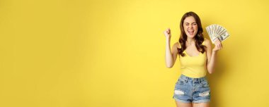 Enthusiastic smiling woman showing money cash, laughing and looking excited, standing in summer clothes against yellow background.