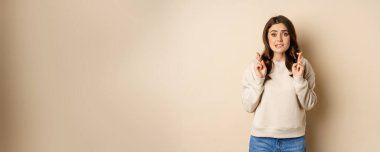 Hopeful worried girl cross fingers, making wish, standing over beige background.