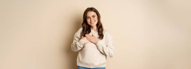 Smiling woman holding hands on heart, looking with care and love at camera, standing in sweater over beige background.