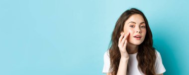 Cilt bakımı. Genç bir kadının aynaya baktığı, parlak cildine dokunduğu, kozmetik ürünlerini kontrol ettiği, mavi arka planda duran, güzellik ürünlerini uyguladığı,.