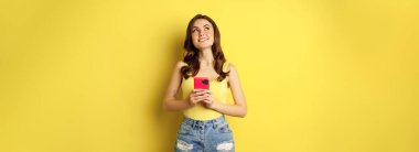 Technology and cellphone. Beautiful stylish girl order in online store, praying in application, using smartphone app and thinking, making decision, standing over yellow background.