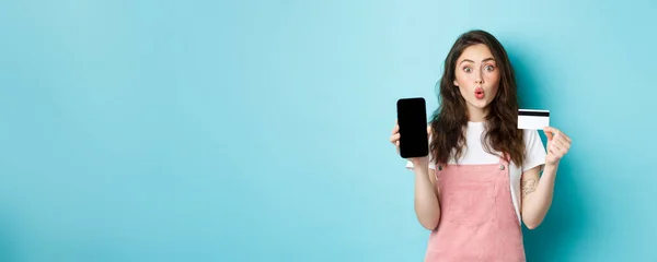 stock image Portrait of young beautiful woman showing plastic credit card and empty smartphone screen, look amused and interested, showing interesting app, standing over blue background.