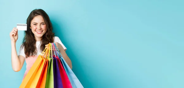 若い満足した女性の笑顔 プラスチッククレジットカードを表示し ショッピングバッグを保持 非接触支払いで購入 青の背景の上に立って — ストック写真