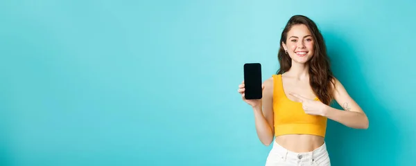 stock image Happy attractive woman showing advertisement on smartphone screen, pointing at empty display of phone and smiling, standing over blue background.