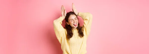 一个风趣可爱的年轻女子的画像 露出舌头 笑容满面 做东兔子的手势 站在粉红的背景下 — 图库照片