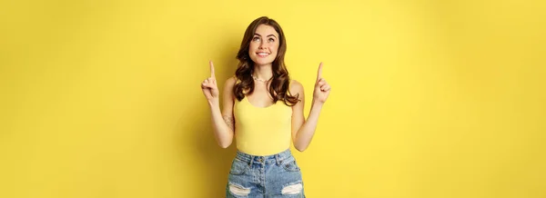 stock image Portrait of cheerful girl smiling, pointing fingers up, showing promo text, summer advertisement, standing against yellow background.