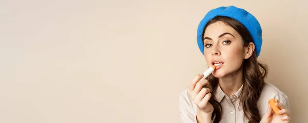 stock image Close up portrait of young beatufiul woman looking in mirror and fixing her makeup, put on lipstick, standing over beige background.