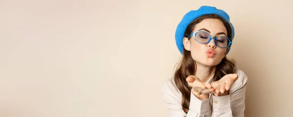 stock image Beautiful stylish woman in sunglasses, blowing air kiss on hands near lips, gazing softly and flirty at camera, posing against beige background.