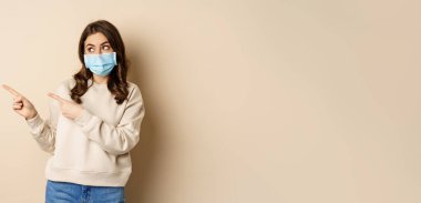 Health and covid-19 concept. Cute brunette woman in medical face mask, pointing fingers left at logo, banner, showing copy space, standing over beige background.