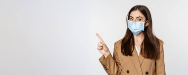 Coronavirus and people concept. Portrait of business woman at workplace wearing face mask, pointing finger left at logo, company banner, white background.