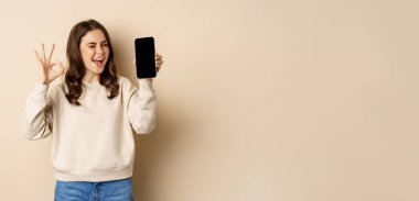 Beautiful modern woman winks, shows okay sign and mobile phone screen, app interfce, standing over beige background.
