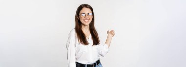 Smiling brunette woman, office lady showing way, pointing behind, demonstrating smth behind her, standing over white background.