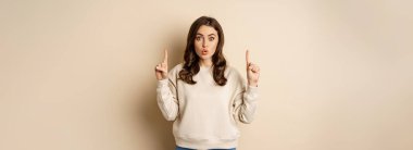 Surprised beautiful girl showing advertisement, pointing fingers on top, store sale or website link up, standing over beige background.