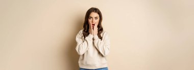 Portrait of shocked brunette woman drop jaw, gasping and staring speechless at camera, beige background. Copy space