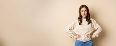 Frustrated, offended young woman looking hurt ad insulted at camera, standing shocked over beige background. People and emotions concept