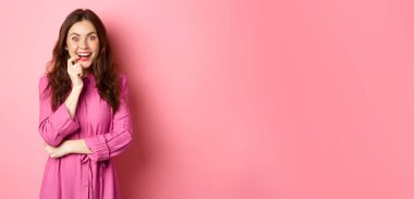 Heyecanlı güzel bir kız gülümsüyor, parmağını ısırıyor ve cezbedici bir yüzle kameraya bakıyor, ilginç bir fikre sahip, ilgi çekici bir şeye bakıyor, pembe arka planda duruyor. Boşluğu kopyala