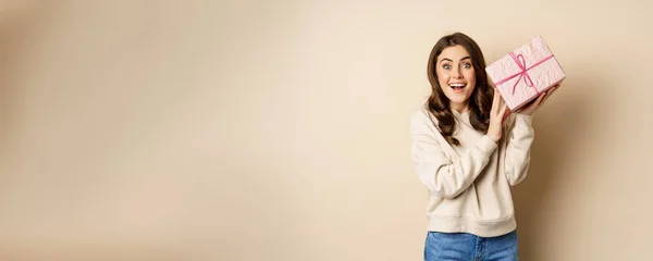 stock image Excited cute girl shaking box with gift, guessing whats inside present, standing happy against beige background.