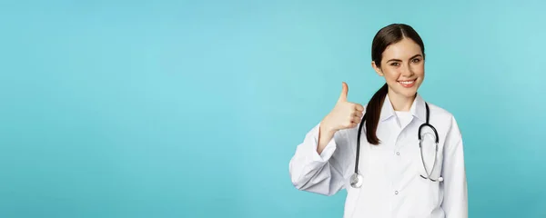 微笑的女医生 带听诊器的保健工作者 在审批时展示大拇指 推荐诊所或药店 站在绿松石背景旁边 — 图库照片