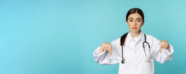 Sad and gloomy doctor, female medical worker pointing fingers down, looking upset, showing bad news, standing in white coat over blue background.