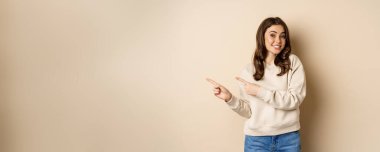 Cheerful attractive female model pointing fingers left, showing store advertisement, banner or logo, standing in sweater over beige background.
