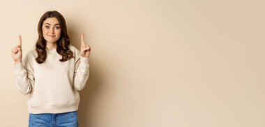 Smiling young woman looking indecisive, shrugging and smirking while pointing fingers up, standing over beige background.