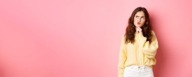 Bilmece gibi tatlı bir kız, düşünceli, dokunaklı çene ve kaşlarını çatmış düşünceli, düşünceli yüzüyle logo afişine bakan, pembe arka planda duran..